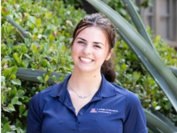 Roarke Poggio, wearing a navy blue University of Arizona College of Education polo, smiles outdoors in front of green bushes and agave plants. She stands with relaxed posture and natural lighting.