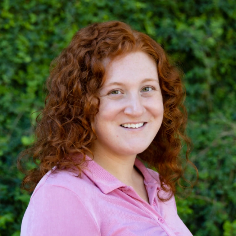 Portrait of Jillian Hanlin smiling outdoors, wearing a pink collared shirt, with curly red hair and a green leafy background.