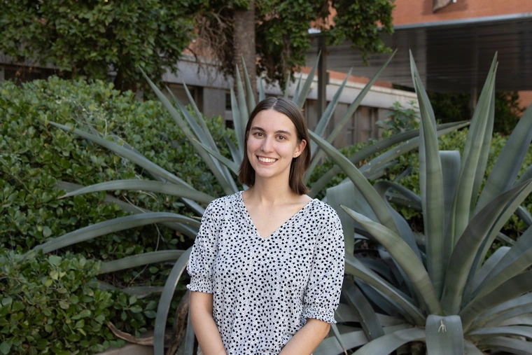 Portrait of Mackenzie Frithsen, a young woman with shoulder-length straight brown hair, smiling and wearing a white blouse with a black floral pattern, standing in front of large green agave plants and bushes.