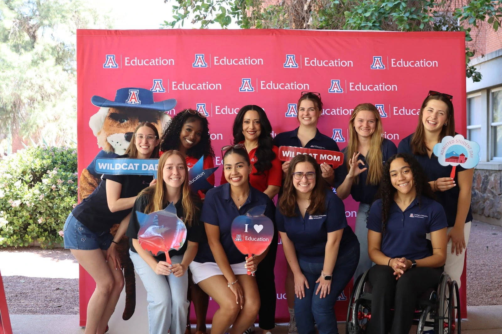 A diverse group of University of Arizona College of Education students and staff pose smiling in front of a red step-and-repeat banner with the College of Education logo. Some hold fun signs including “Wildcat Dad,” “Wildcat Mom,” and “Admitted Student Day,” while Wilbur Wildcat's cutout appears on the left.